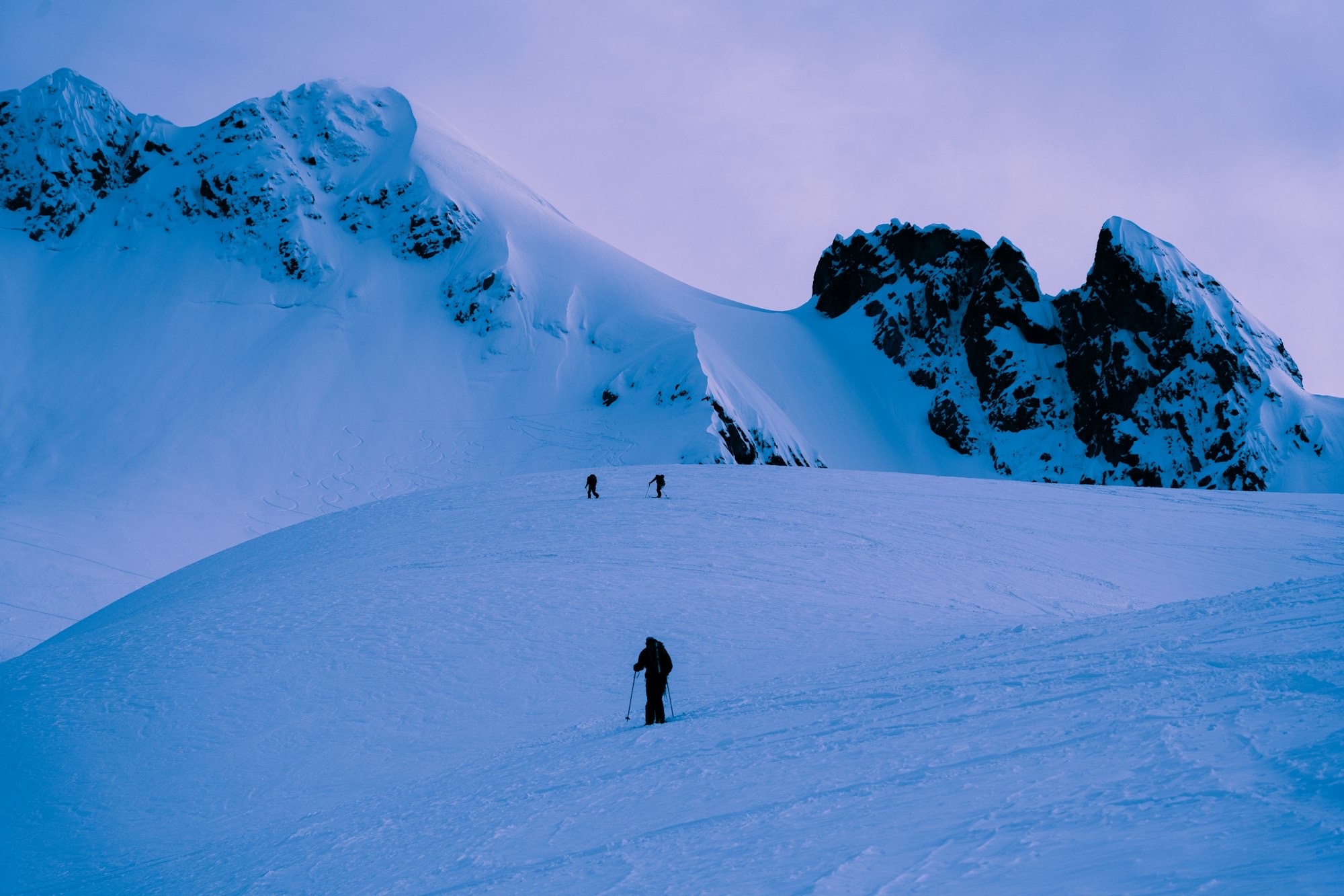 Ski tourers heading toward a jagged rock ridge in blue dusk light