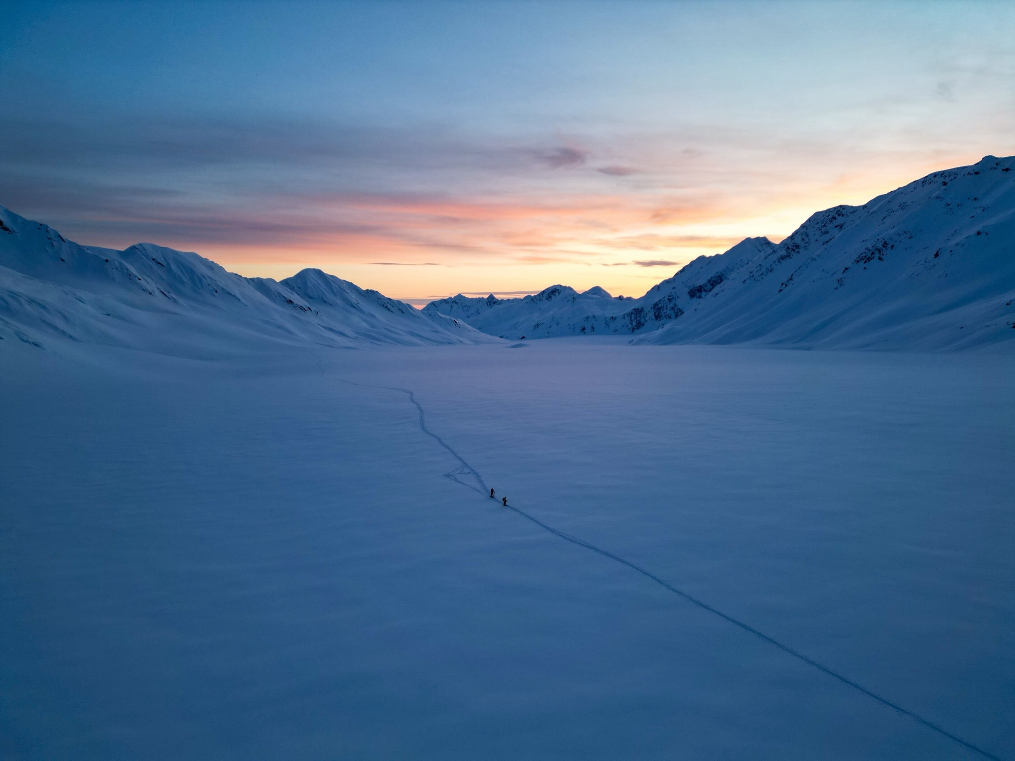 Two skiers on a long skin track across a snowy valley at sunset